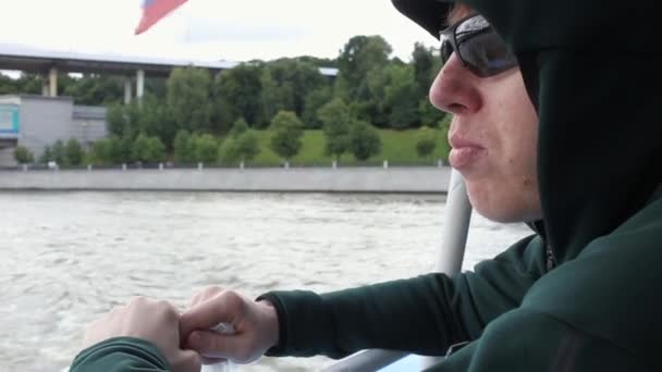 Primer plano del joven en gafas de sol y capucha, masticando contra el agua, flotando en el río en barco. viaje de transporte marítimo — Vídeos de Stock