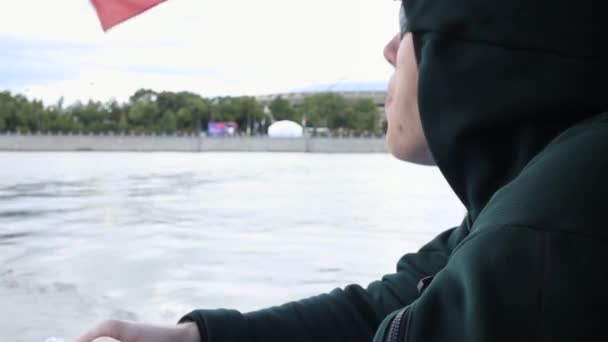 Hombre en gafas de sol, capucha, come semillas, escupe, navega en el río en barco. viaje de transporte acuático. ondeando bandera rusa — Vídeos de Stock