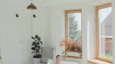 Spor giyimli esmer genç kız lotus pozisyonunda kapalı gözlerle oturur ve evdeki parlak odada meditasyondan zevk alır. Sağlıklı bir yaşam tarzı. Yoga yapmak, stres atmak, kafa dinlemek, dinlenme odası.