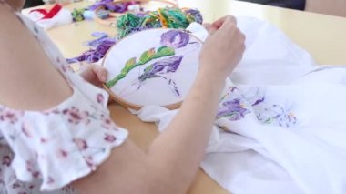 Yazlık elbiseli genç kız kafeterya masasında oturmuş iğneyle dikiş dikiyor. Nakışlı halka, renkli desenli nakışlı kumaş. Geleneksel hobi, yaşam tarzı. İğne işi, el işi.