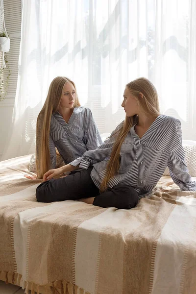 Dos hermanas gemelas bonitas en jeans negros y camisas a rayas con el pelo largo sentado en el sofá contra el fondo de la ventana — Foto de Stock