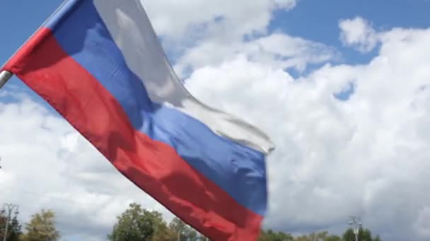 俄罗斯国旗在绿树和城市手电筒的映衬下迎风飘扬 阳光灿烂的日子里 蓝蓝的天空笼罩着白云 — 图库视频影像