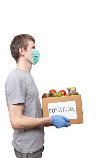 volunteer in medicine mask, protective gloves holding grocery food in carton donation box: vegetable oil, grape, tin cans, tomatoes. Coronavirus relief funds, charity help, delivery, local support