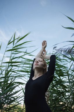 Siyah elbiseli, uzun, ıslak sarı saçlı, kolları havada, yeşil kamışların karşısında duran, şehvetli, genç, beyaz bir kızın portresi. Doğada poz veren çekici bir kadın.