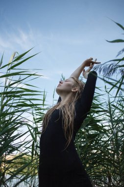 Siyah elbiseli, uzun, ıslak sarı saçlı, kolları havada, yeşil kamışların karşısında duran, şehvetli, genç, beyaz bir kızın portresi. Doğada poz veren çekici bir kadın.