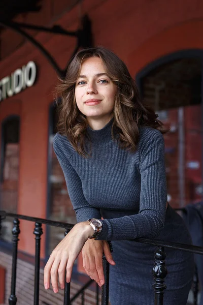 Gri balıkçı yakalı, kolunda saatleri olan, kırmızı binanın önünde poz veren genç, düşünceli, güzel bir kız. Çekici bir kadının sokak fotoğrafı, zarif bir modelin şehir fotoğrafı.