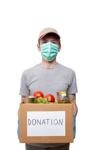 volunteer in medicine mask, protective gloves holding grocery food in carton donation box: vegetable oil, grape, tin cans, tomatoes. Coronavirus relief funds, charity help, delivery, local support