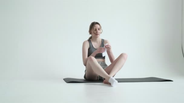 Szczupła dziewczyna w odzieży sportowej siada na macie na białym cykloramie, zakłada skarpetki na nogi — Wideo stockowe