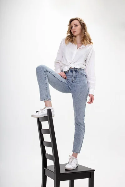 Jovem menina branca pensiva posa em camisa branca, jeans azul na cadeira no estúdio — Fotografia de Stock