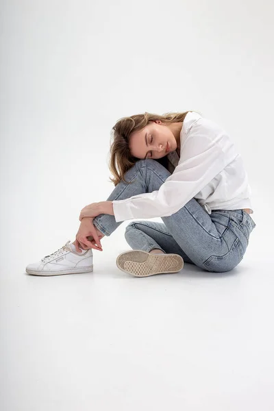 Young cute pensive caucasian girl posing in white shirt, blue jeans at studio — Stock Photo, Image