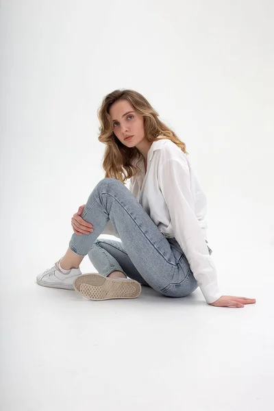 Молода мила стомлена біла дівчина позує в білій сорочці, блакитні джинси в студії — стокове фото