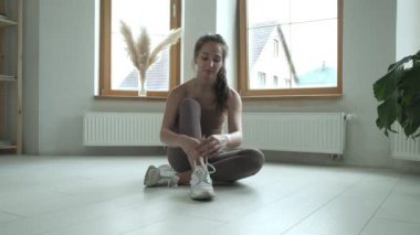 Spor giysili genç bir kız dizlerinin üzerinde oturuyor, ayakkabı bağcıklarını spor ayakkabılarına bağlıyor.