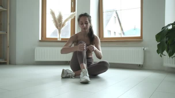 Spor giysili genç bir kız dizlerinin üzerinde oturuyor, ayakkabı bağcıklarını spor ayakkabılarına bağlıyor. — Stok video