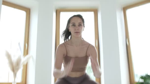 운동복을 입은 젊은 갈색 머리 소녀는 점프를 하고 밝은 방에 앉아 집에서 — 비디오