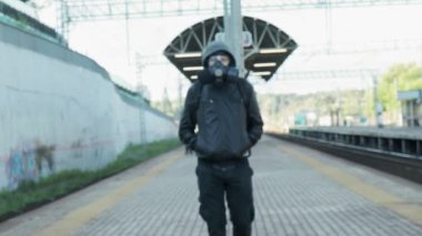 Gaz maskeli, kapüşonlu ve sırt çantalı bir adam demiryolu platformunda yürüyor.
