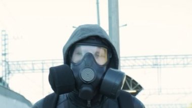 Gaz maskeli adam, kapüşonlu ceket hareket halindeki trenin önünde yürüyor.