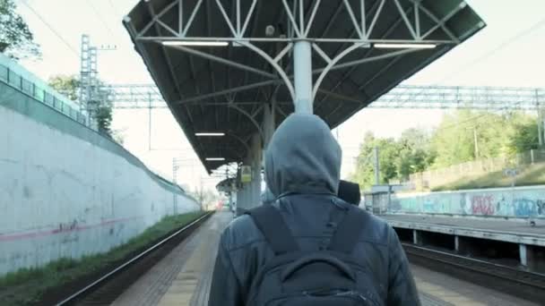 Назад человека в противогазе с капюшоном и рюкзаком ходьба по железнодорожной платформе — стоковое видео