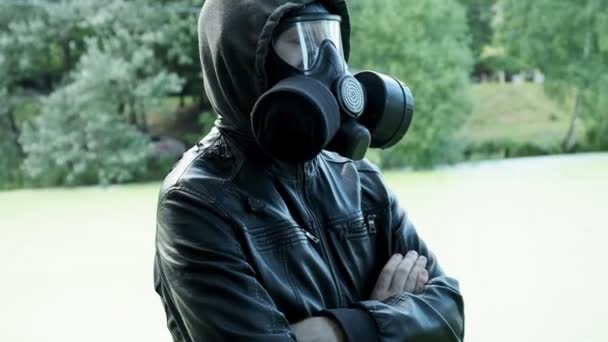 Людина в газовій масці, шкіряна куртка з капюшоном, що стоїть на відкритому повітрі в лісі біля озера — стокове відео