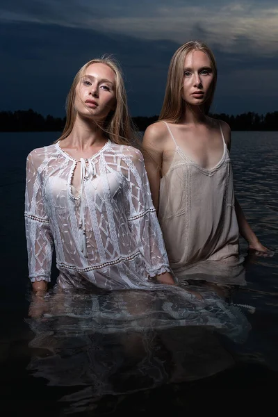 Δύο νεαρές δίδυμες αδελφές ποζάρουν με ελαφρά φορέματα στο νερό της λίμνης το βράδυ του καλοκαιριού — Φωτογραφία Αρχείου
