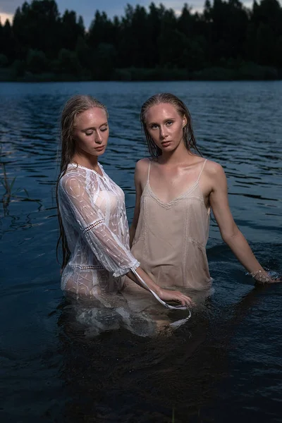 Deux jeunes sœurs jumelles posant en robes claires dans l'eau du lac la nuit d'été — Photo