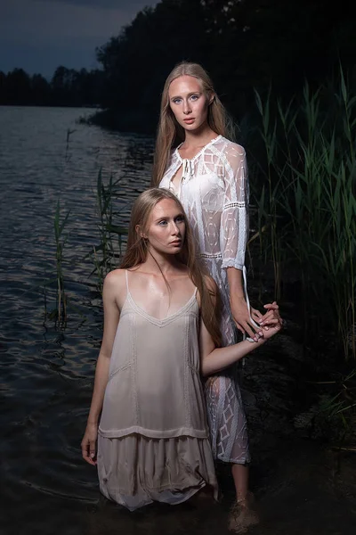 Deux jeunes sœurs jumelles posant en robes claires dans l'eau du lac la nuit d'été — Photo