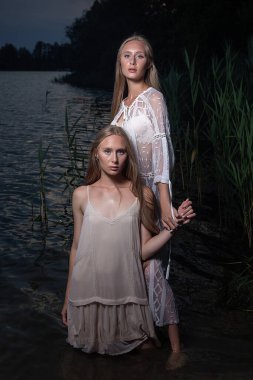 İki genç ikiz kız kardeş yaz gecesi gölün sularında açık renk elbiseler giyip poz veriyorlar.