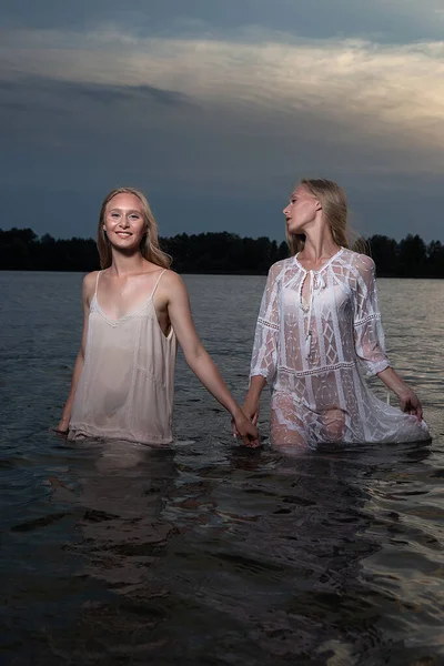 Deux jeunes sœurs jumelles posant en robes claires dans l'eau du lac la nuit d'été — Photo