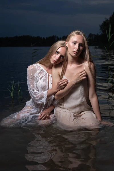 two young twin sisters posing in light dresses in water of lake at summer night