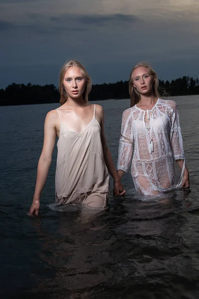 Дві молоді сестри-близнюки позують у легких сукнях у воді озера в літню ніч — стокове фото