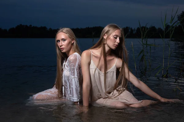 Deux jeunes sœurs jumelles posant en robes claires dans l'eau du lac la nuit d'été — Photo