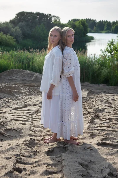Dos hermanas gemelas jóvenes atractivas posando en la playa de arena en ropa blanca elegante — Foto de Stock