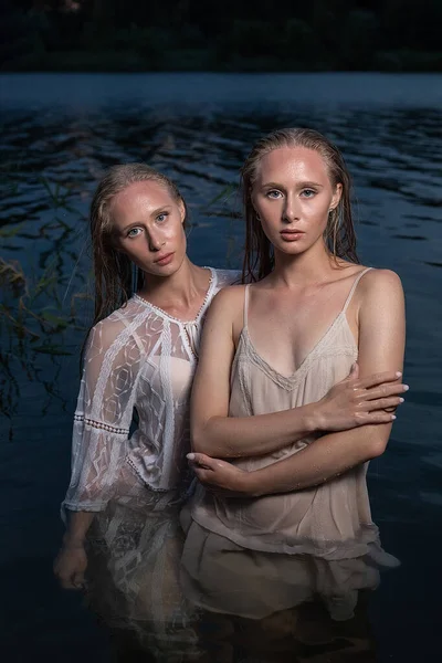 Δύο νεαρές δίδυμες αδελφές ποζάρουν με ελαφρά φορέματα στο νερό της λίμνης το βράδυ του καλοκαιριού — Φωτογραφία Αρχείου