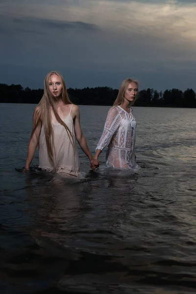 Deux jeunes sœurs jumelles posant en robes claires dans l'eau du lac la nuit d'été — Photo