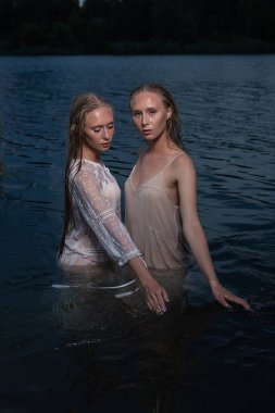 İki genç ikiz kız kardeş yaz gecesi gölün sularında açık renk elbiseler giyip poz veriyorlar.