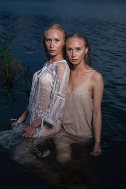 İki genç ikiz kız kardeş yaz gecesi gölün sularında açık renk elbiseler giyip poz veriyorlar.