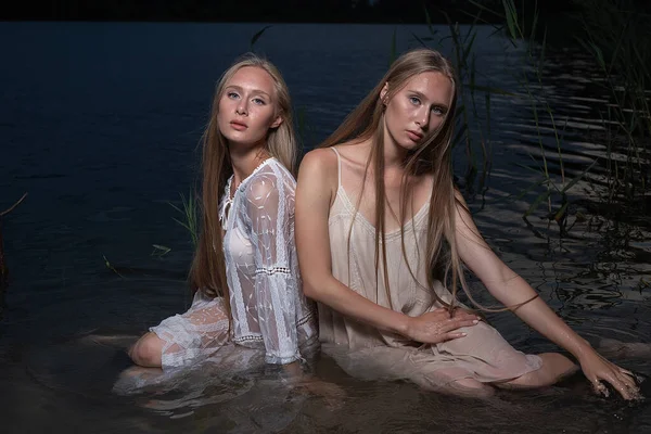 Deux jeunes sœurs jumelles posant en robes claires dans l'eau du lac la nuit d'été — Photo