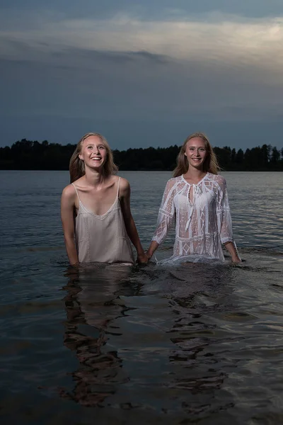 Dos hermanas gemelas jóvenes posando en vestidos ligeros en el agua del lago en la noche de verano —  Fotos de Stock