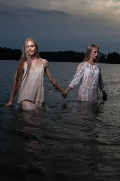 Twee jonge tweeling zussen poseren in licht jurken in het water van het meer in de zomer nacht — Stockfoto