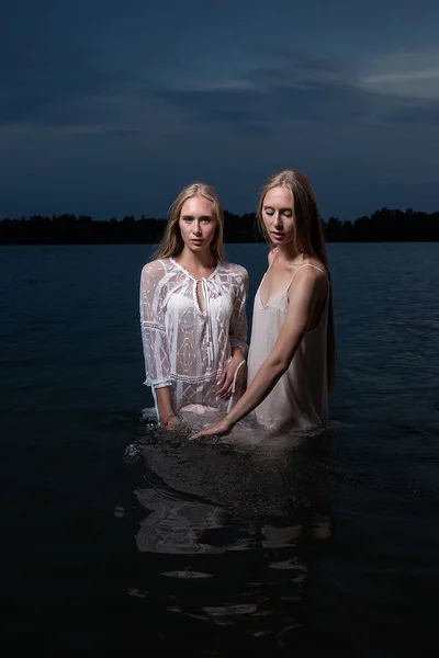 Deux jeunes sœurs jumelles posant en robes claires dans l'eau du lac la nuit d'été — Photo