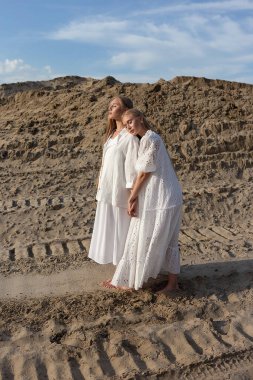 Kum madeninde zarif beyaz giysiler içinde poz veren iki çekici genç kız kardeş.