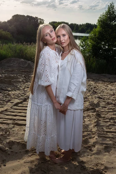 Молоді близнюки з довгим світлим волоссям позує на піщаному пляжі в елегантному білому одязі — стокове фото