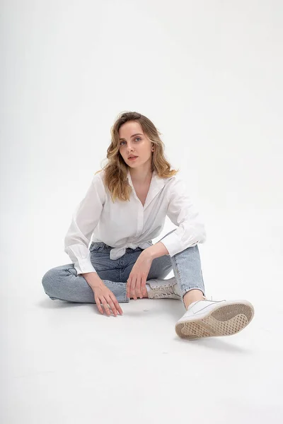 Mujer caucásica posando en camisa y jeans azules, sentado en el piso del estudio blanco — Foto de Stock