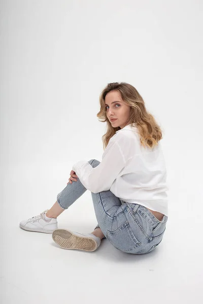Біла жінка позує в сорочці і блакитних джинсах, сидячи на білій підлозі студії — стокове фото