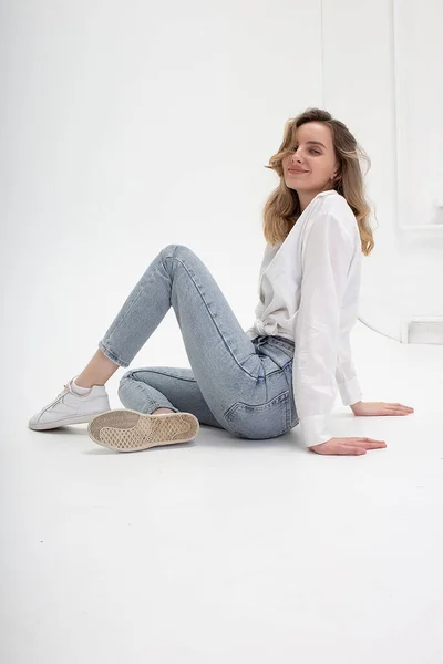 Caucasian woman posing in shirt and blue jeans, sitting on white studio floor — Stock Photo, Image