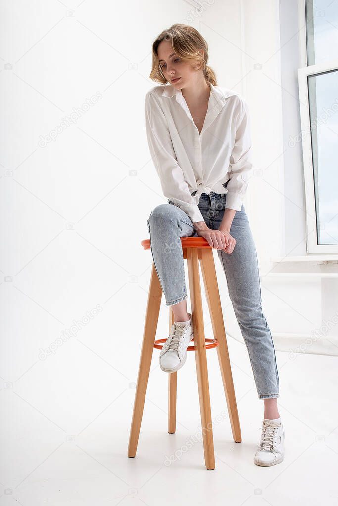 young pensive caucasian woman posing in shirt and blue jeans, sitting on stool