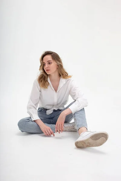 Mujer caucásica posando en camisa y jeans azules, sentado en el piso del estudio blanco — Foto de Stock