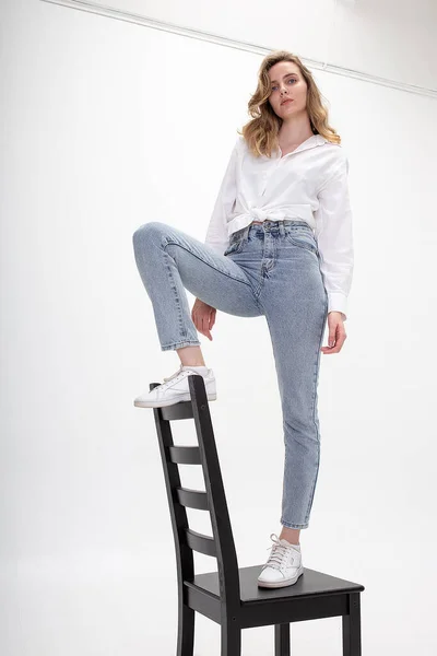 Giovane ragazza caucasica pensosa posa in camicia bianca, jeans blu sulla sedia in studio — Foto Stock