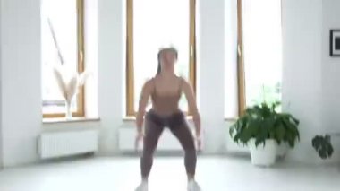 Spor giysili genç esmer kız atlıyor, evdeki aydınlık odada çömeliyor.