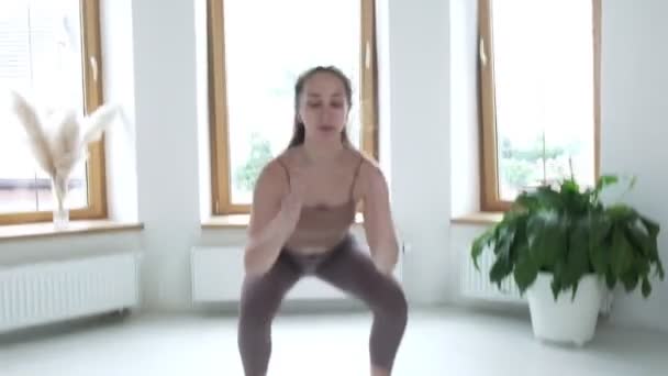 Młoda brunetka w odzieży sportowej skacze, przysiada w jasnym pokoju w domu — Wideo stockowe
