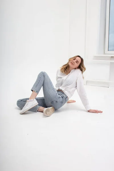 Kaukasiska kvinna poserar i skjorta och blå jeans, sitter på vit studio golv — Stockfoto